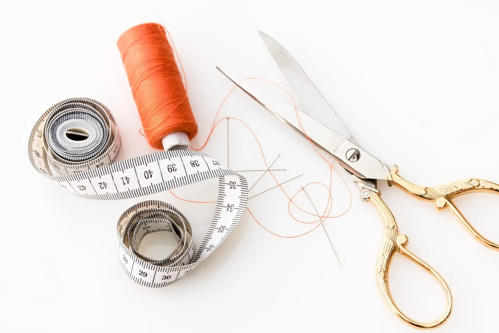 A soft measuring tape with centimetre markings, an orange spool of thread, needles and scissors laid out on a white surface